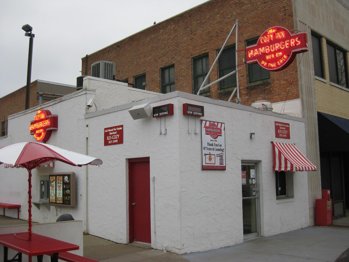 The Cozy Inn building at 108 N 7th Street, Salina, Kansas — a small white stucco storefront with red door and the iconic Cozy Inn Hamburgers neon sign towering above it